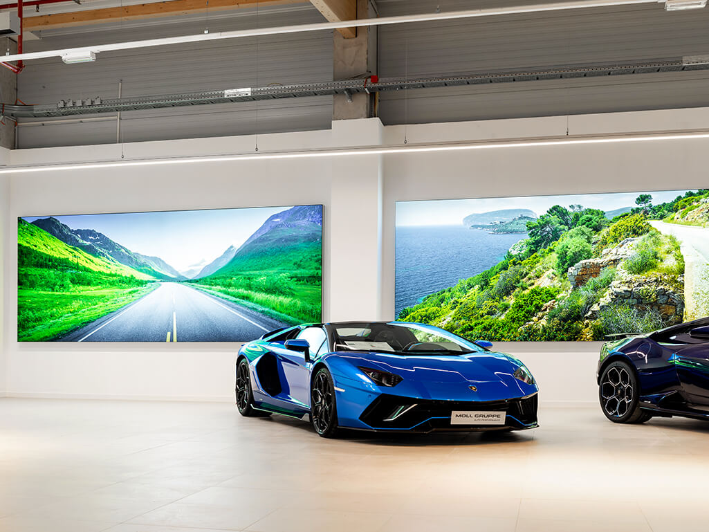 Selbstleuchtende, großformatige Wandbilder mit italienischen Küstenlandschaften in einem Autohaus. Im Vordergrund sind edle italienische Wagen der Marke Lamborghini zu sehen.